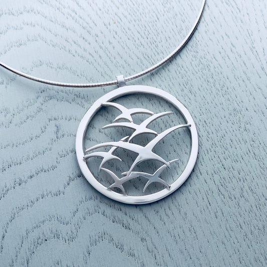 A silver necklet on a wire with a large pendant featuring a flock of flying birds in a round frame
