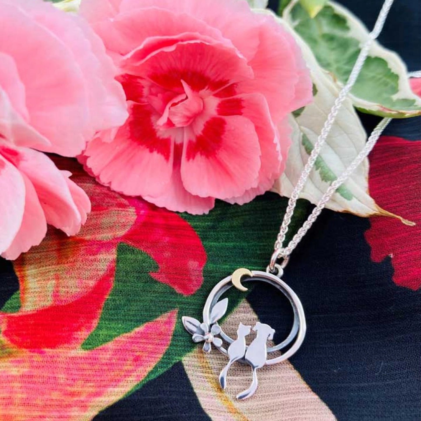 A silver pendant featuring a cat and dog sat together under a gold moon