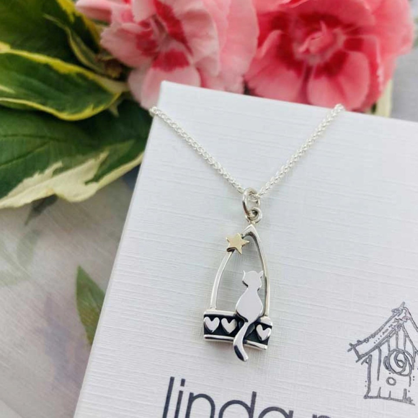 A silver stirrup pendant with a little cat, heart details and a gold star on box