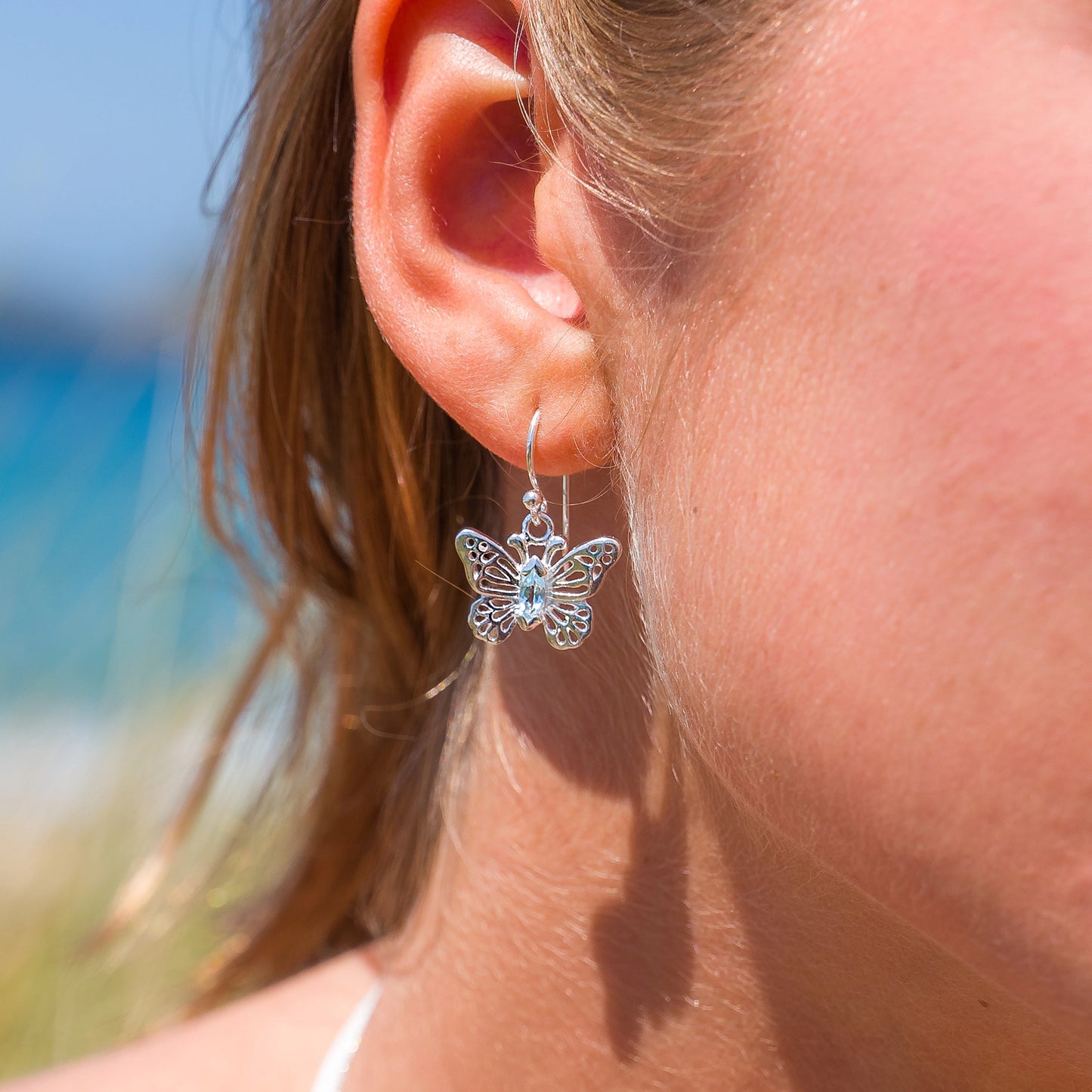 Model wearing silver butterfly drop earrings with filigree details and blue topaz stone int he centre