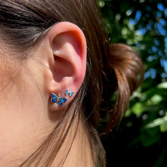 Model wearing a pair of tiny and large blue enamel butterfly stud earrings in polished silver