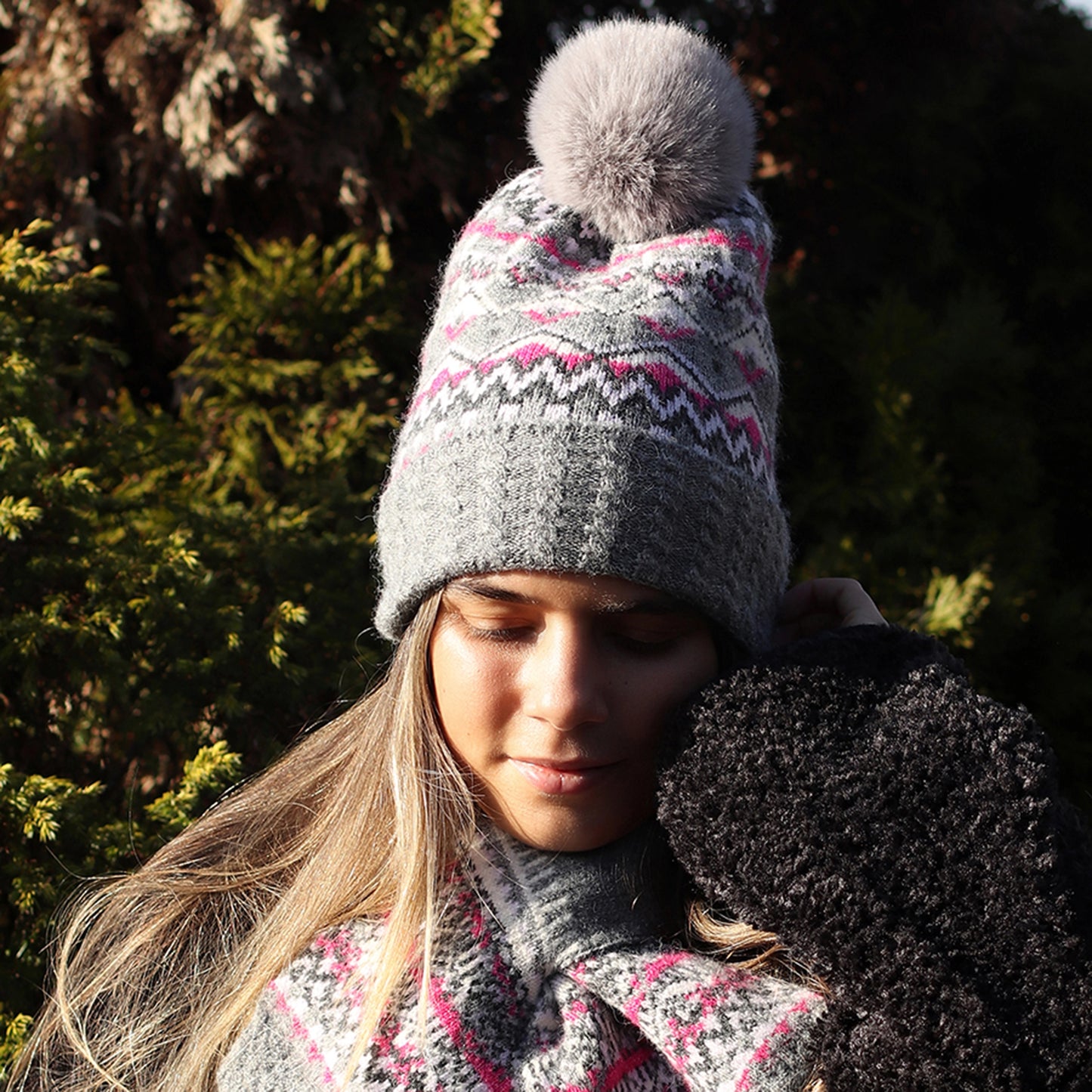 Model wearing knitted fair isle patterned hat in grey and pink with faux fur pompom and cable knit turn up