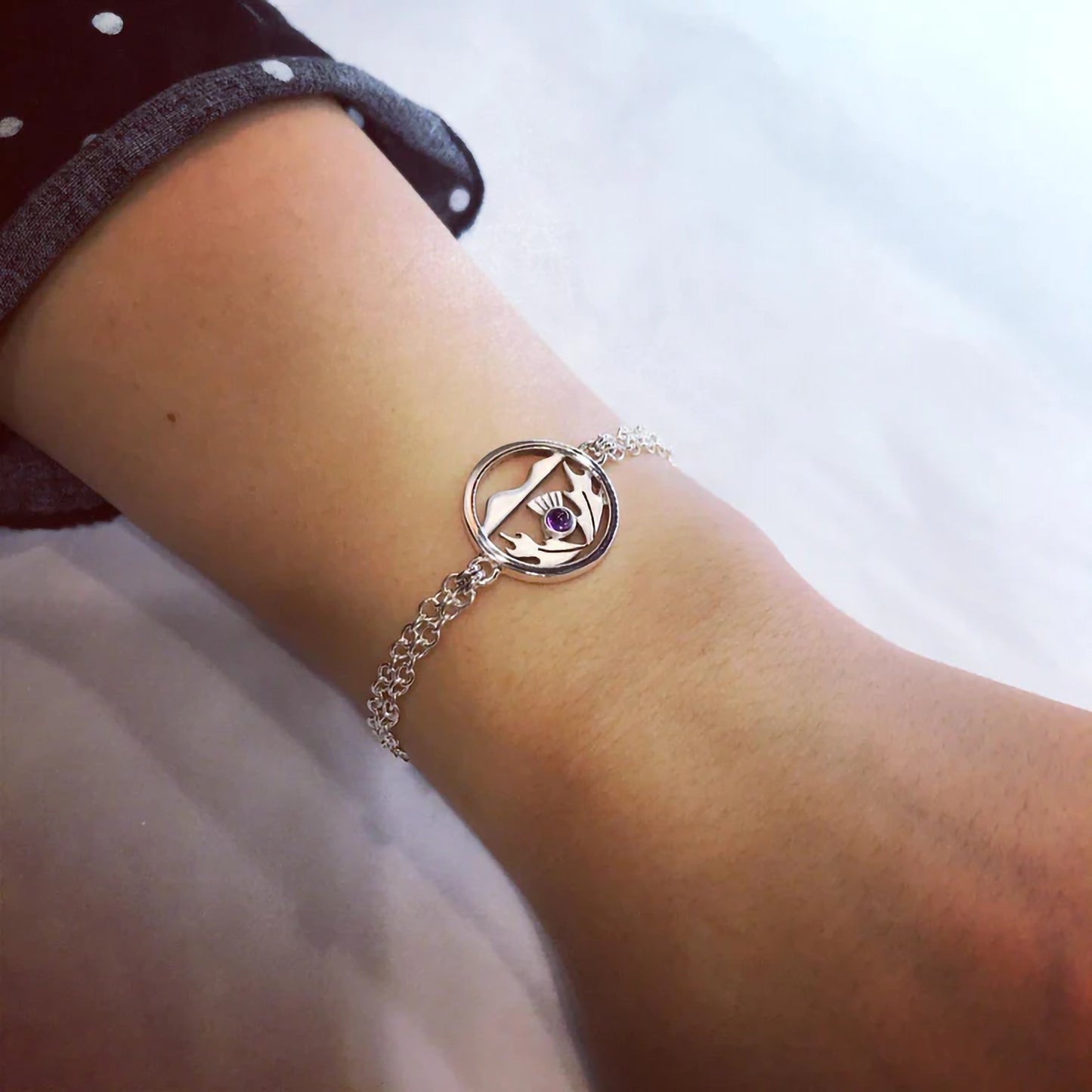 A silver bracelet with a round pendant with a Scottish thistle in front of hills and an amethyst stone