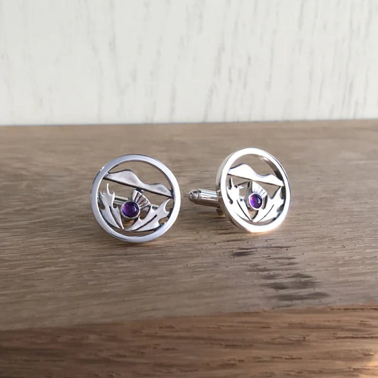 A pair of round silver cufflinks featuring a Scottish thistle with hills in the background and an amethyst stone