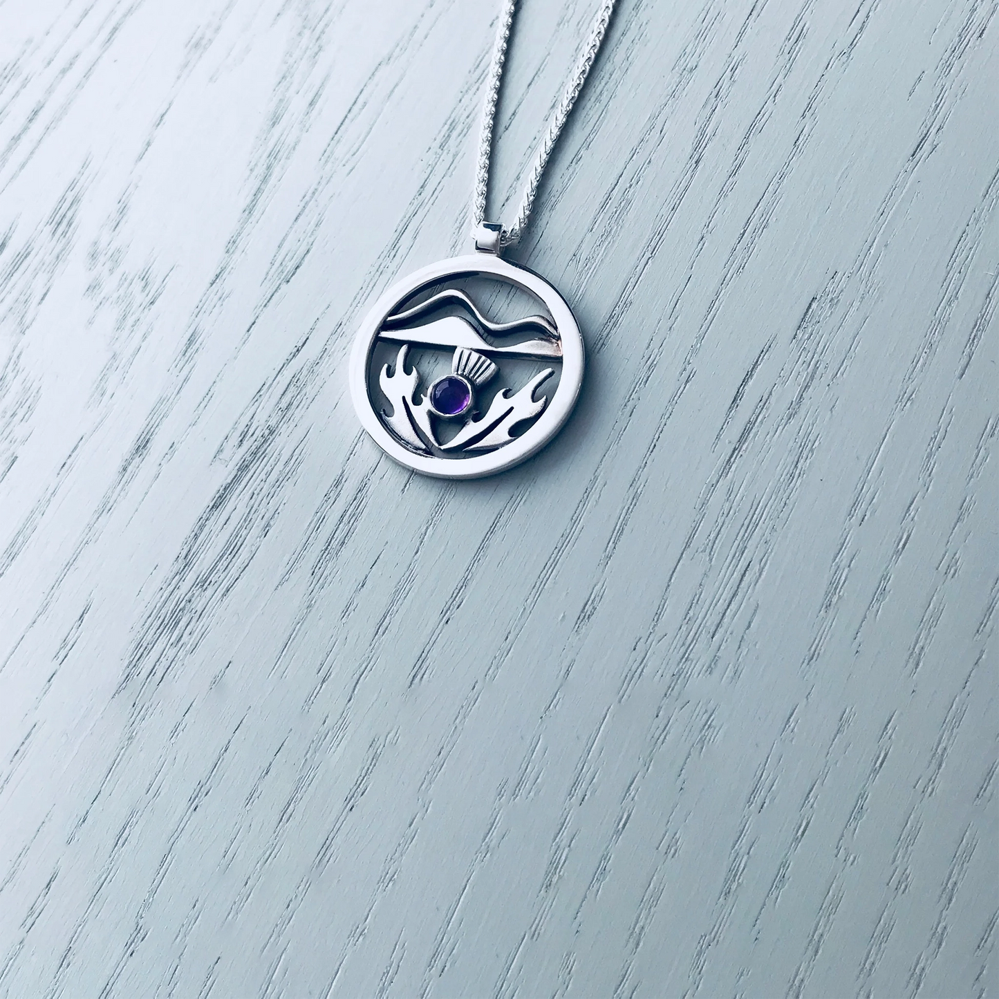 A round silver pendant with a Scottish thistle in front of hills and a purple amethyst stone