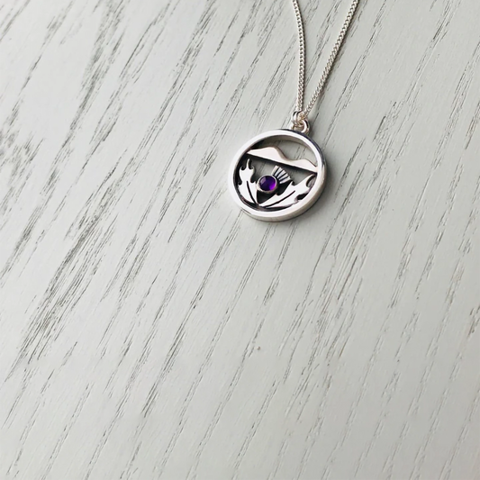 A silver necklace with a pendant featuring a Scottish thistle in front of hill and a purple amethyst stone