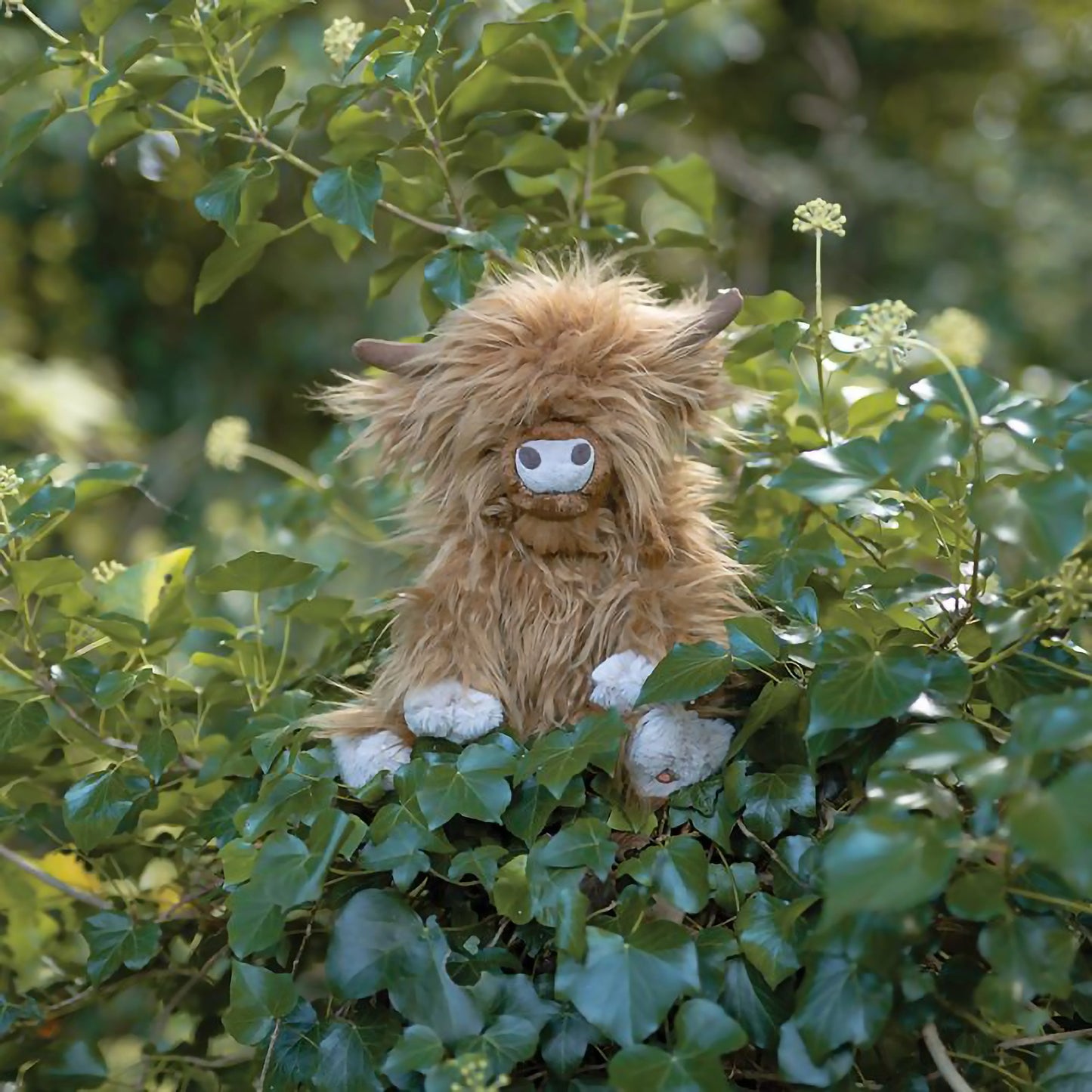 A stuffed Highland cow plush toy with the Wrendale logo embroidered on the bottom of its foot posed in ivy