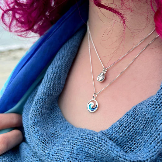 Model wearing silver round pendant with blue enamel ocean wave design and a silver groatie shell necklace