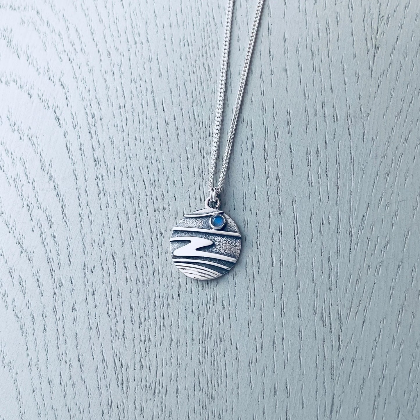 A silver pendant with a round shape featuring raised ripple textures, oxidised silver and a small round moonstone