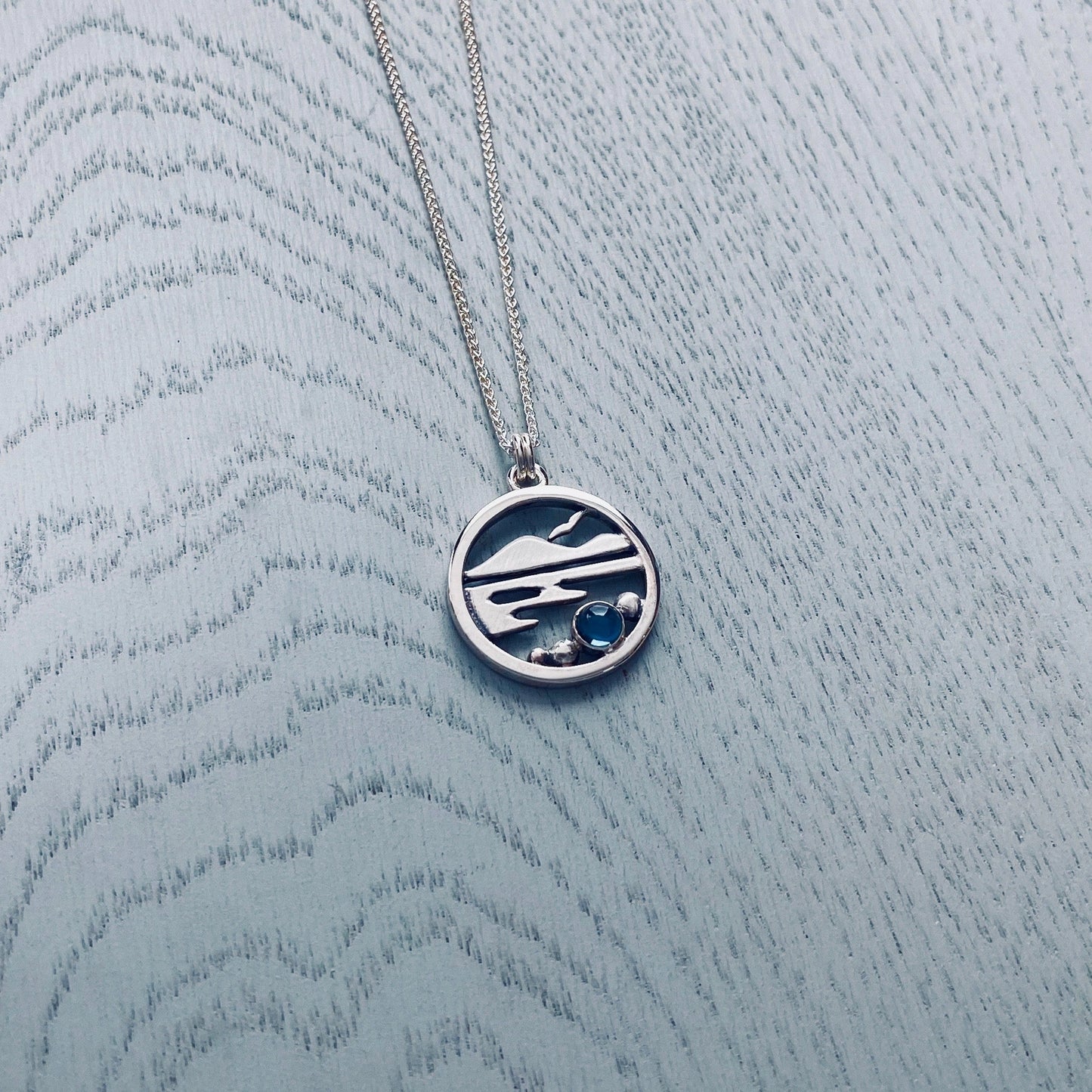 Round silver pendant featuring a Highland scene with Munros, water, a bird, and a blue topaz stone