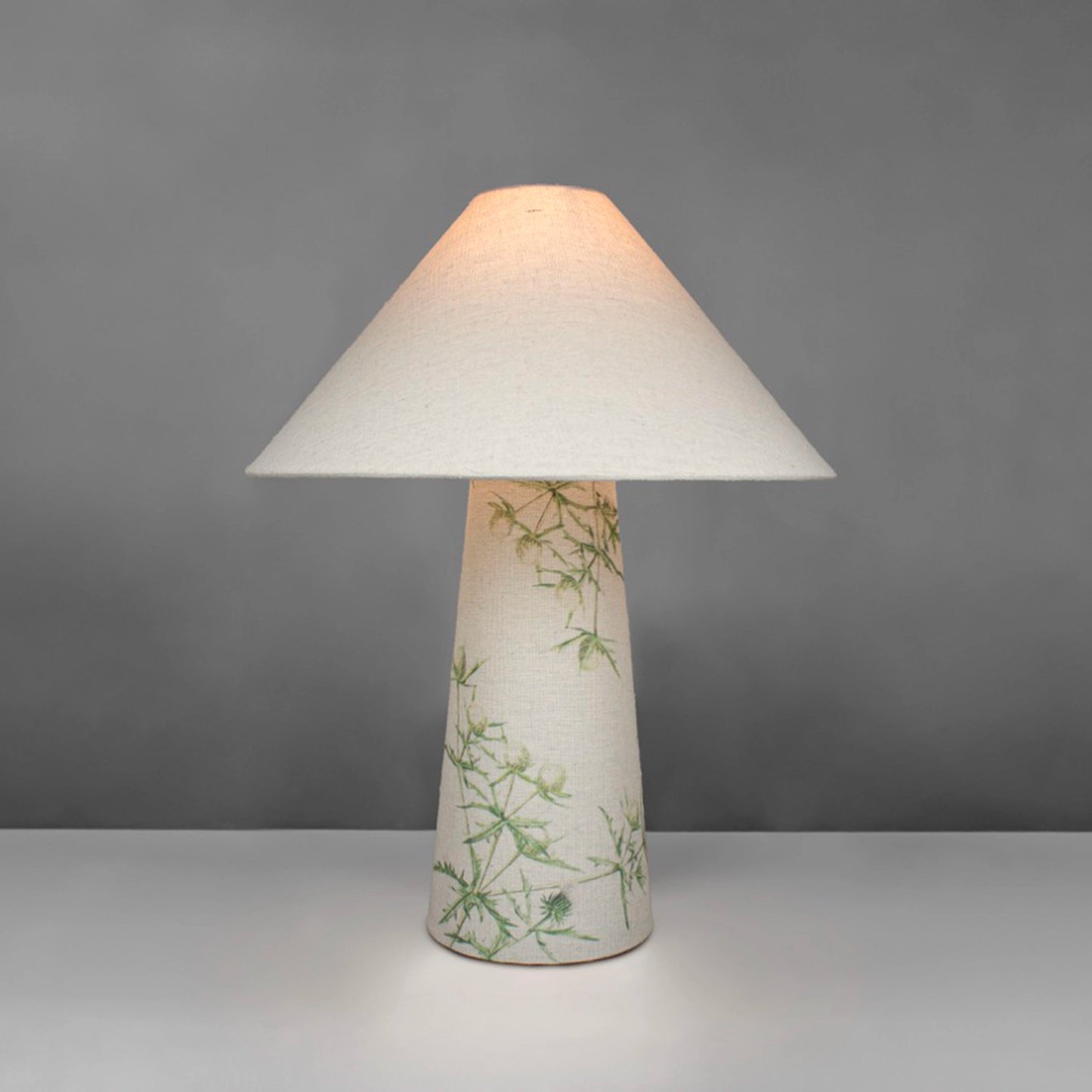 A fabric table lamp with shade and a thistle design on the conical base - lit up