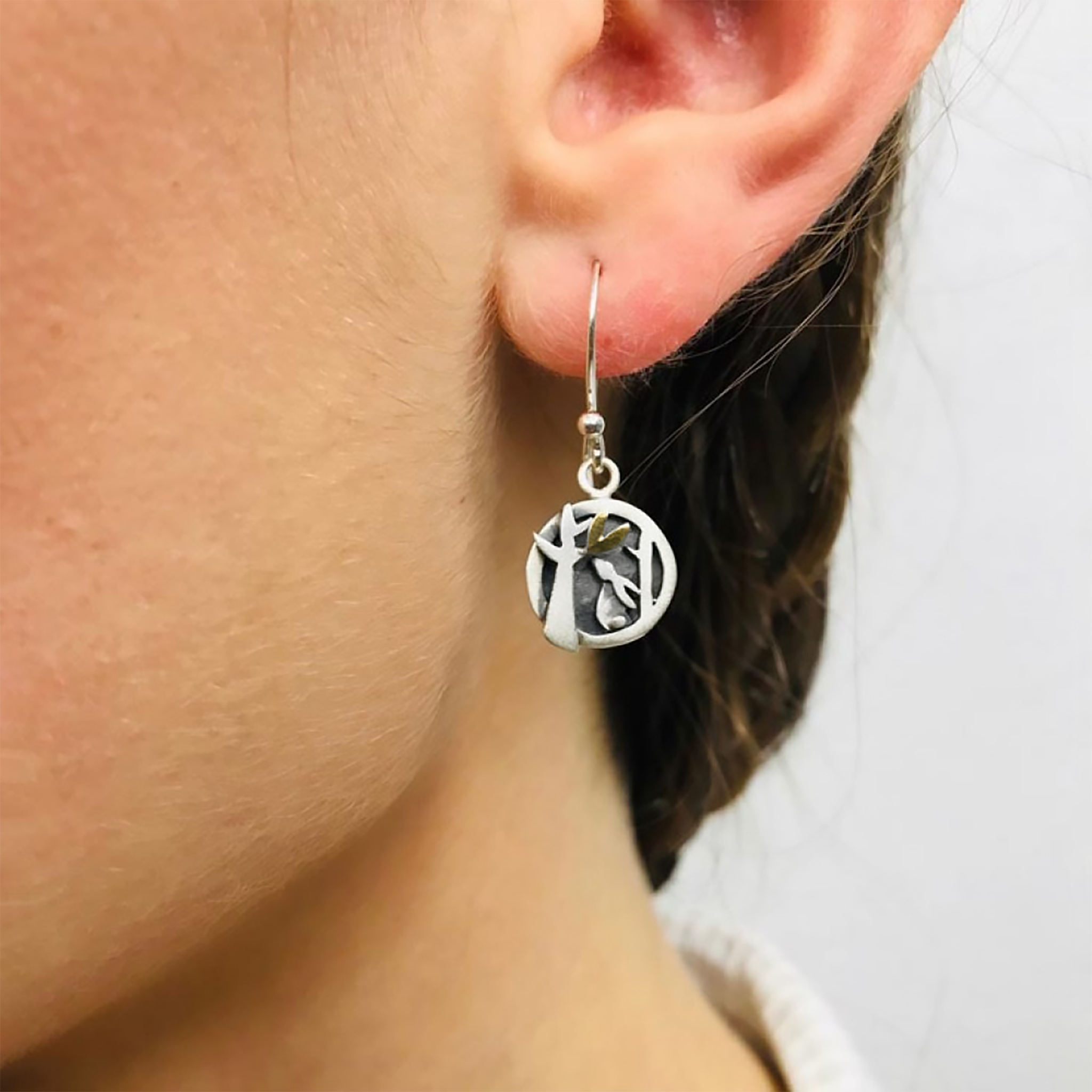 Model wearing pair of round silver earrings with bunny and tree details and a little gold heart on hook fittings