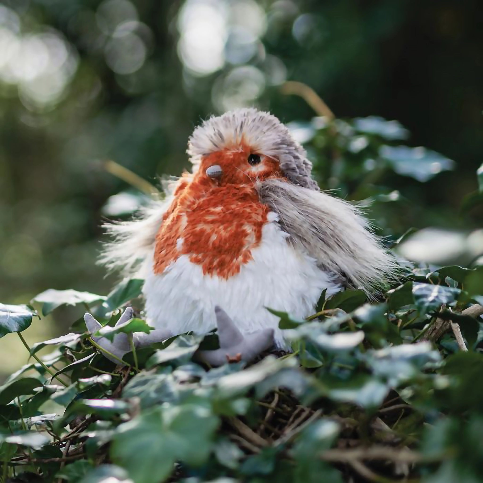 A stuffed robin bird plush toy with the Wrendale logo embroidered on the bottom of its foot posed in ivy