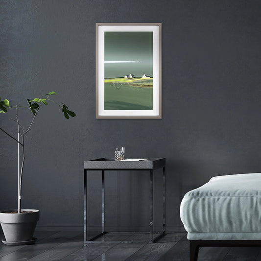 Framed print of a coastal scene in greens and blues with three white cottages on the horizon hanging in a dark room.