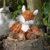 A stuffed fox plush toy with the Wrendale logo embroidered on the bottom of its foot posed on a log