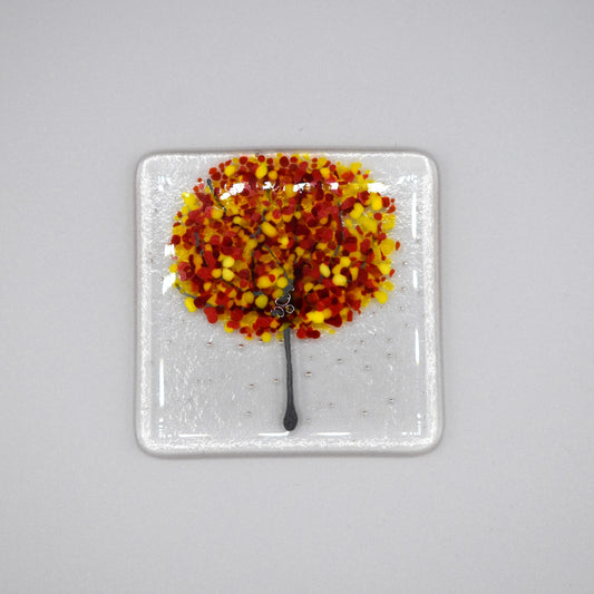 A clear glass square coaster with a yellow & red tree design 