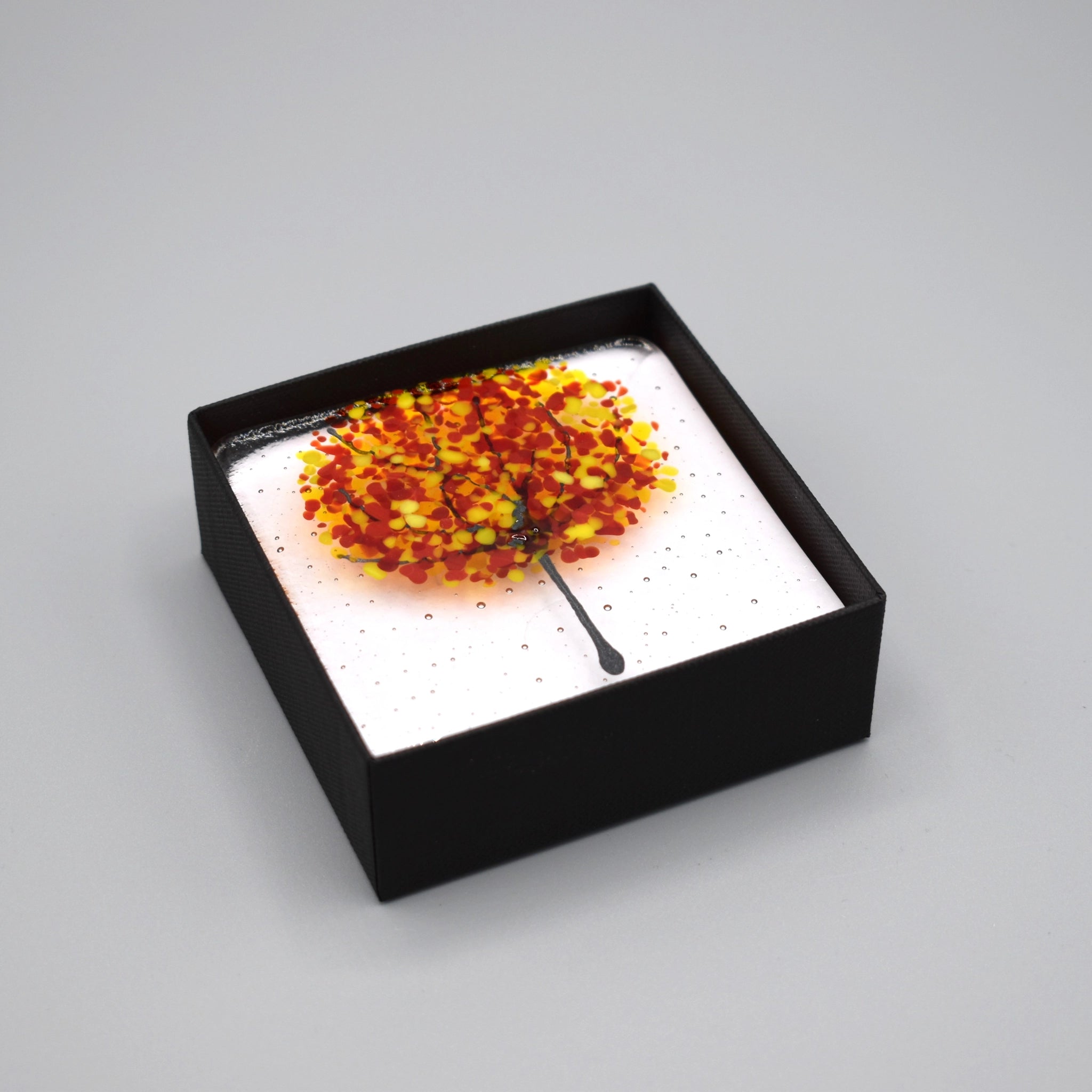 A clear glass square coaster with a yellow & red tree design boxed