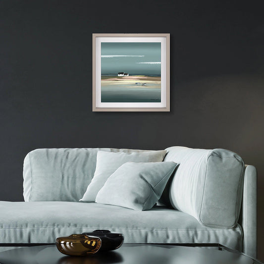Square framed print of a seaside scene with blue water and a white cottage on a sandy shoreline hanging in a dark room.