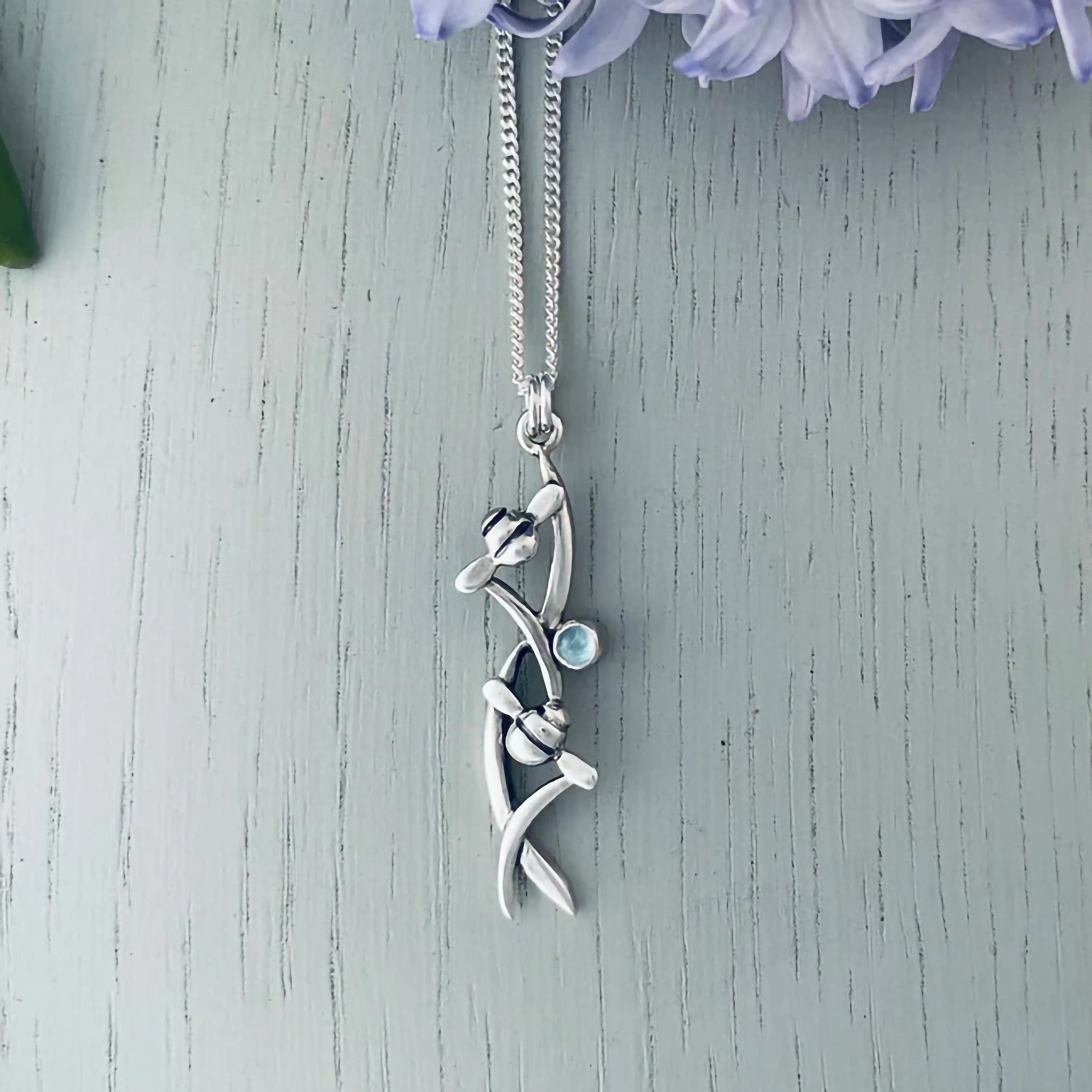 Silver necklace featuring a pendant with two bees in tall grass and a round blue topaz stone