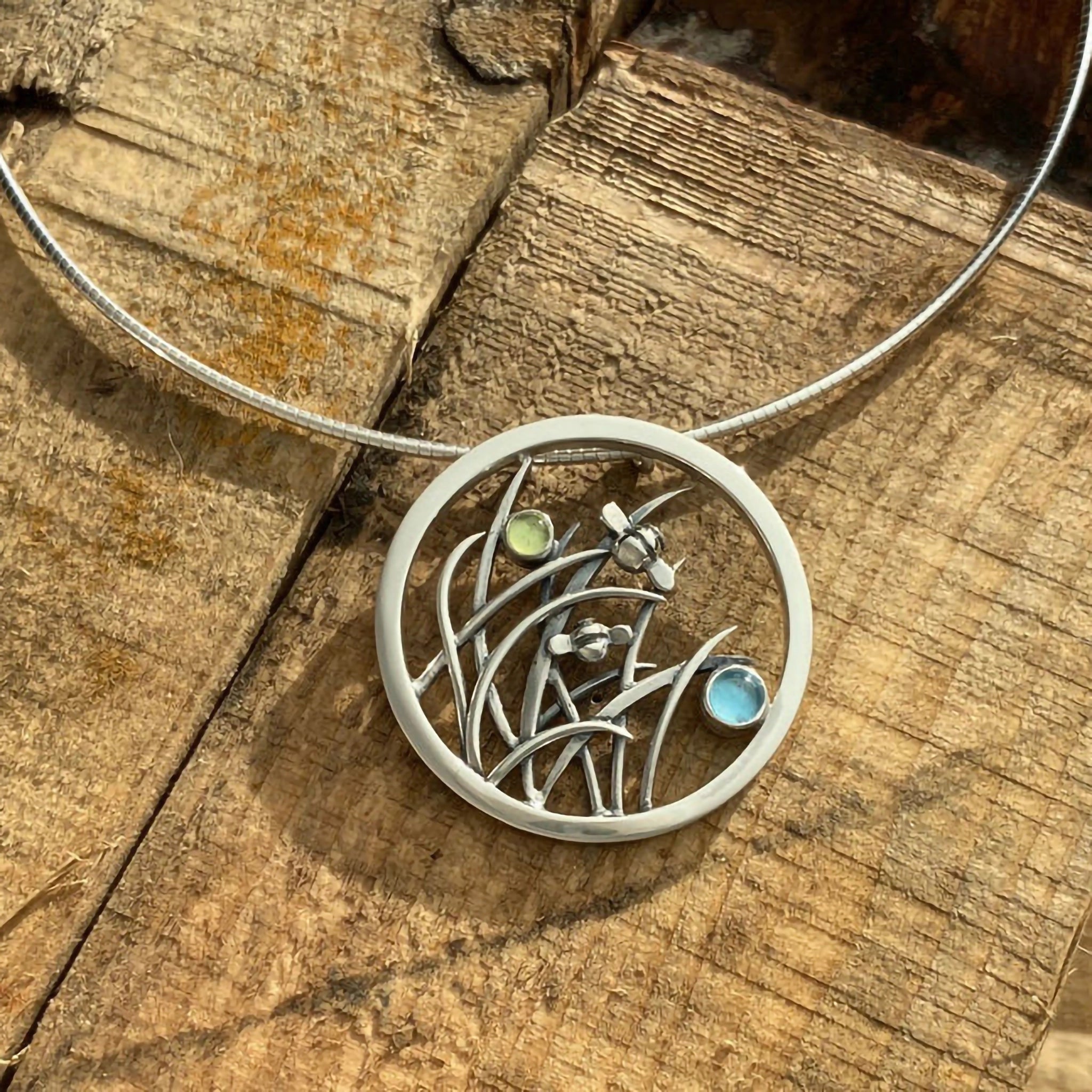 A round necklet featuring bees in tall grass and peridot and blue topaz stones