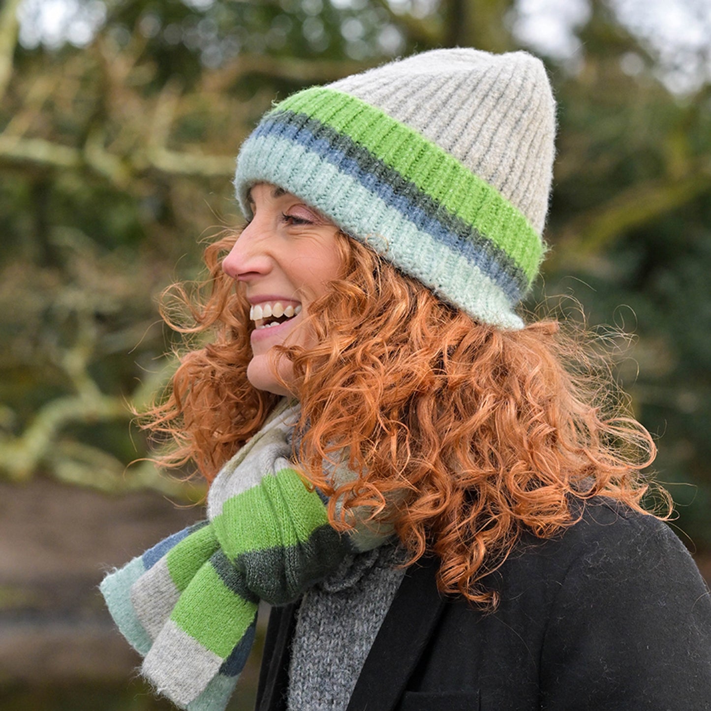 Model wearing grey knitted beanie hat with green and blue striped ribbed turn-up and matching scarf