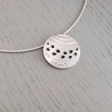 A silver necklet featuring a round pendant with a matt texture, ripple effect and row of cut-out bird prints
