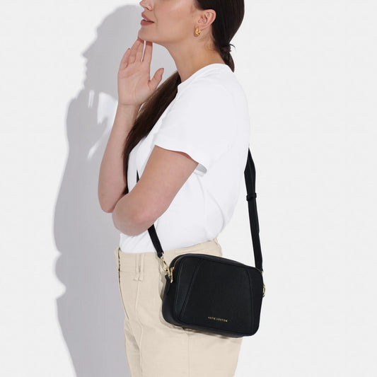 Model wearing a black crossbody bag in a simple box shape with adjustable strap and gold hardware