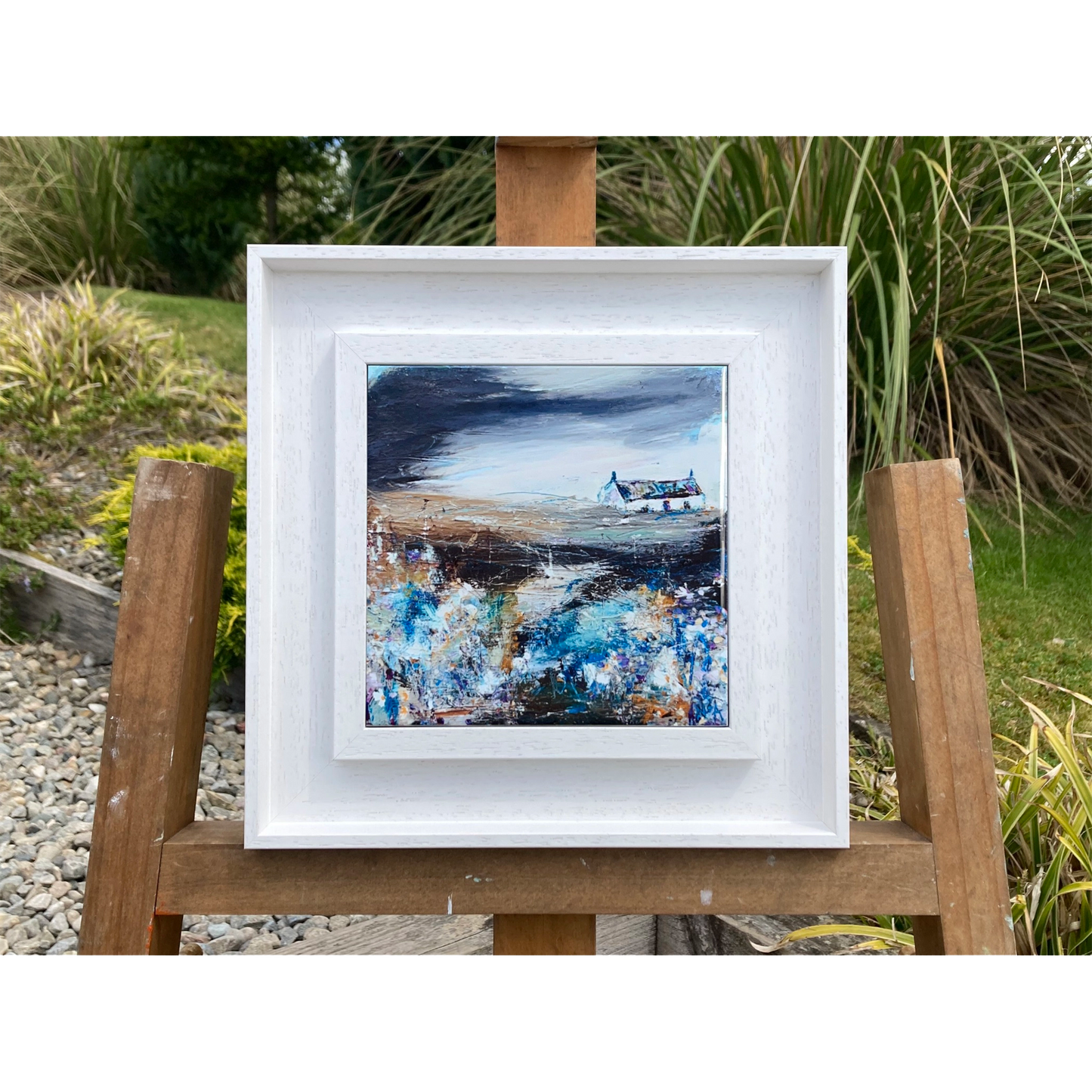 A ceramic tile printed with an abstract painting of a cottage and flowers mounted on a white frame