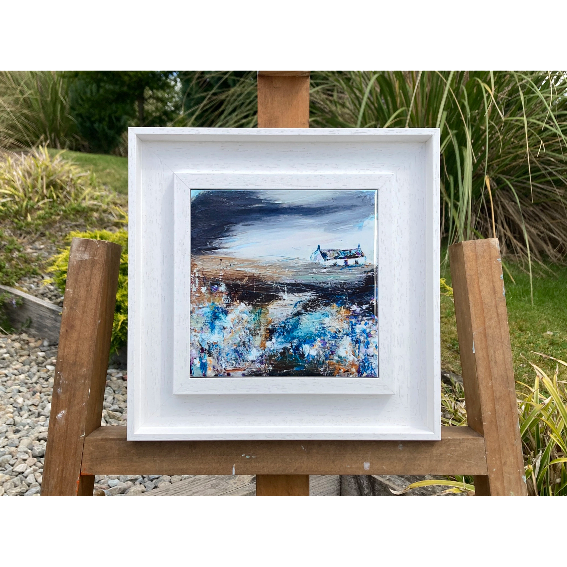 A ceramic tile printed with an abstract painting of a cottage and flowers mounted on a white frame