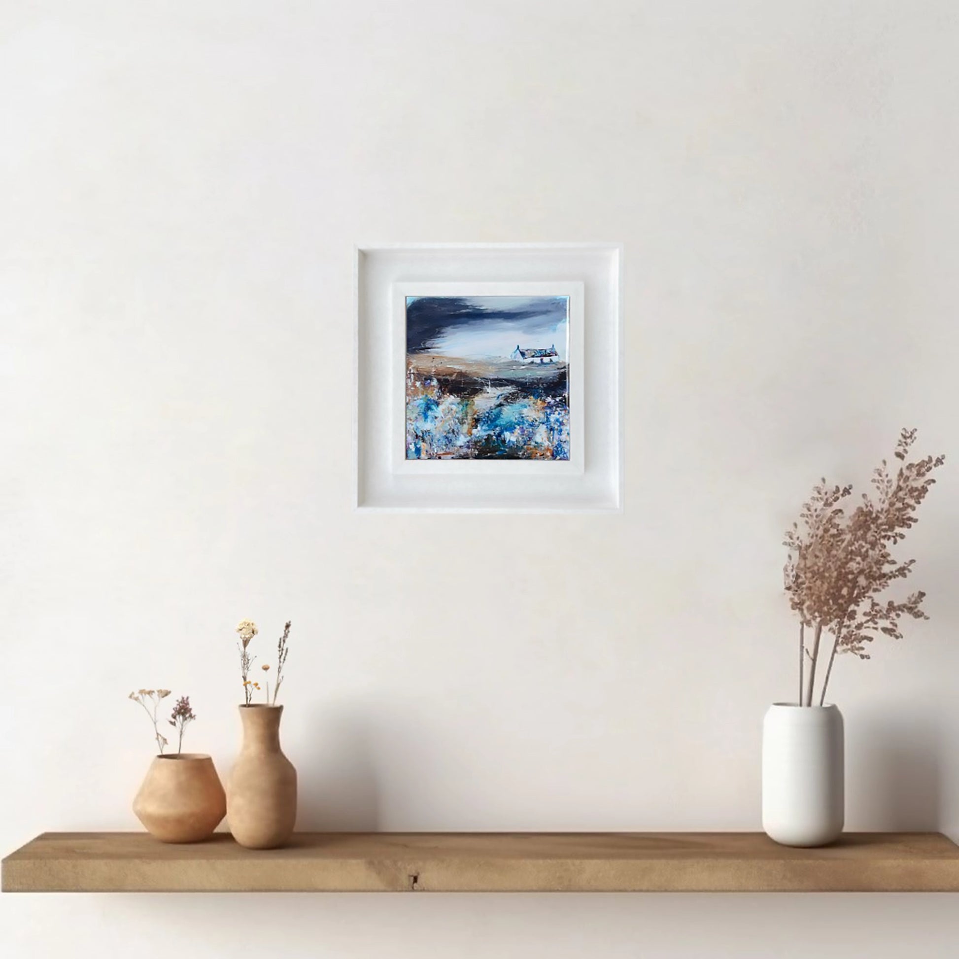 A ceramic tile printed with an abstract painting of a cottage and flowers mounted on a white frame hanging