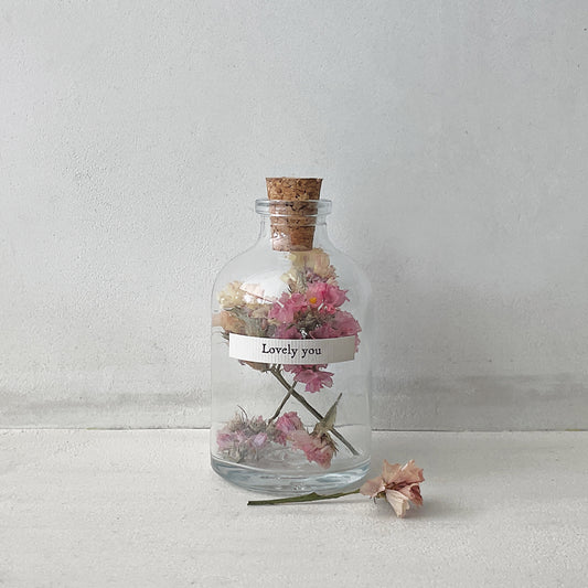 A small cork stopped glass bottle with dried flowers inside and a label that reads 'lovely you'