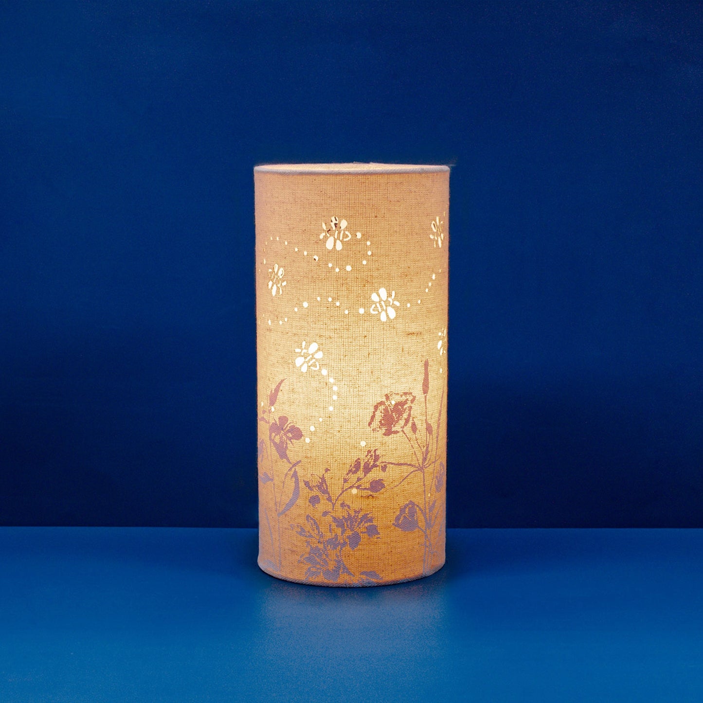 A linen look fabric table lamp with a flower and bumblebee design and cut out leaf details lit up