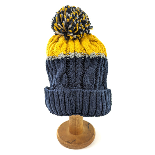 A pompom cable knit hat in navy with mustard top and a grey stripe