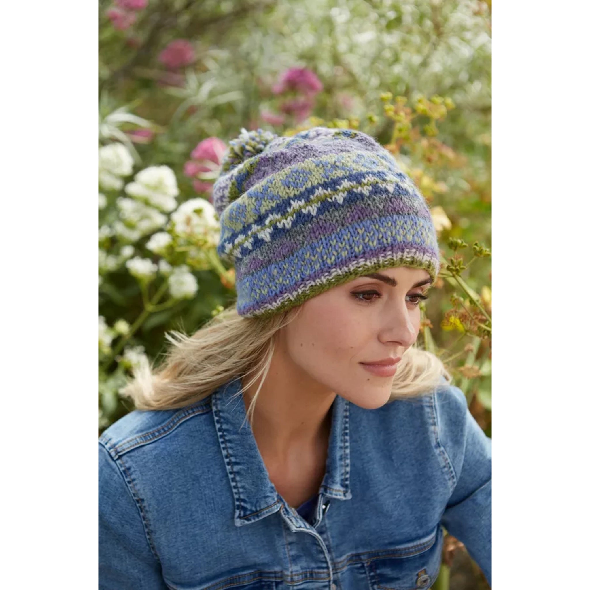 Model wearing a beanie hat with a fairisle pattern in green, purple and blue, and a pompom