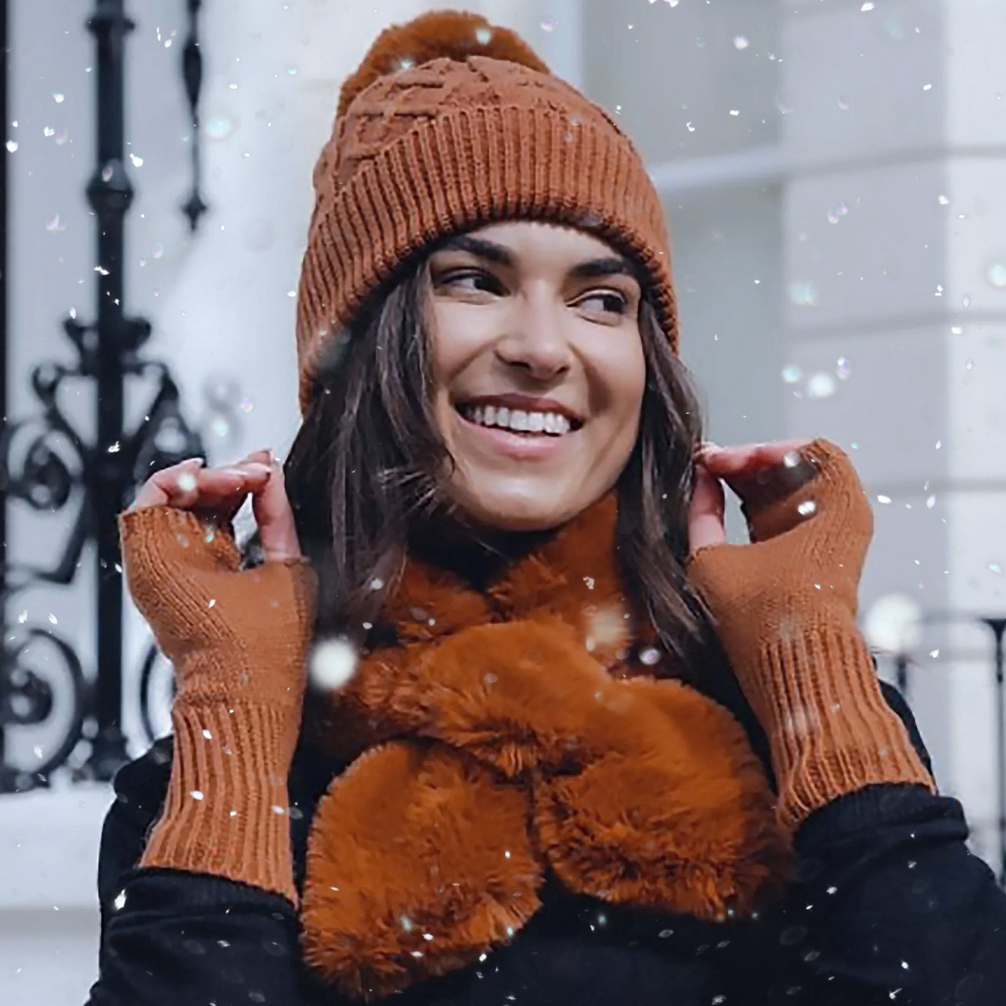 Model wearing nutmeg brown pompom coloured hat, fingerless gloves, and faux fur scarf under snowfall