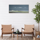  Long framed print of a coastal scene in greens and blues with three white cottages on the horizon hanging in a light room.