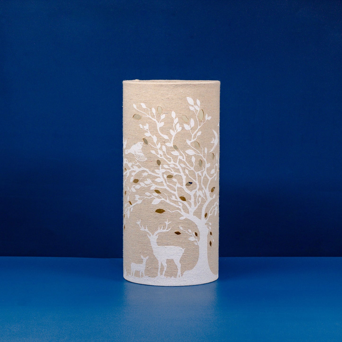 A linen look fabric table lamp with a tree and deer design and cut out leaf details