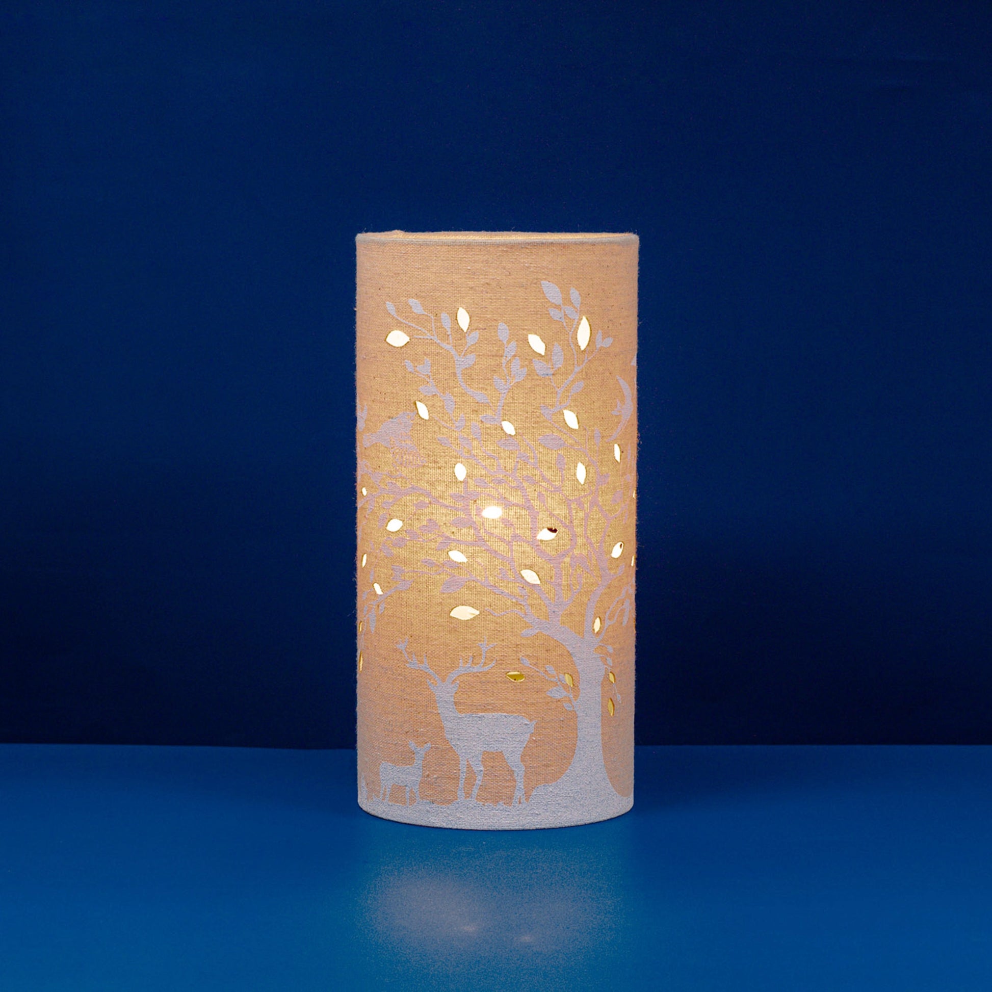 A linen look fabric table lamp with a tree and deer design and cut out leaf details lit up