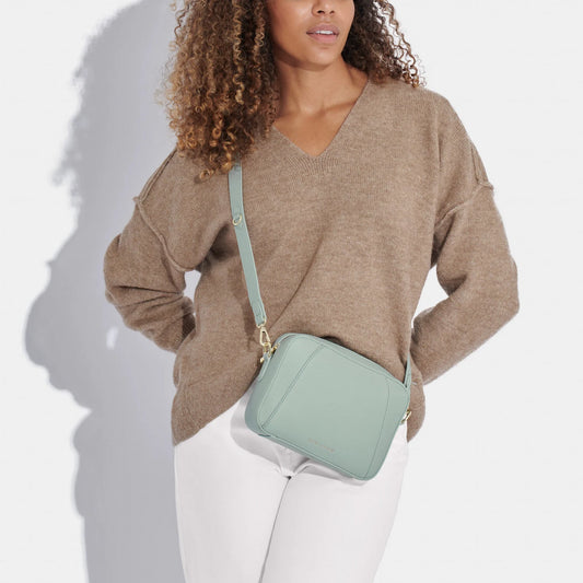 Model wearing a light blue crossbody bag in a simple box shape with adjustable strap and gold hardware