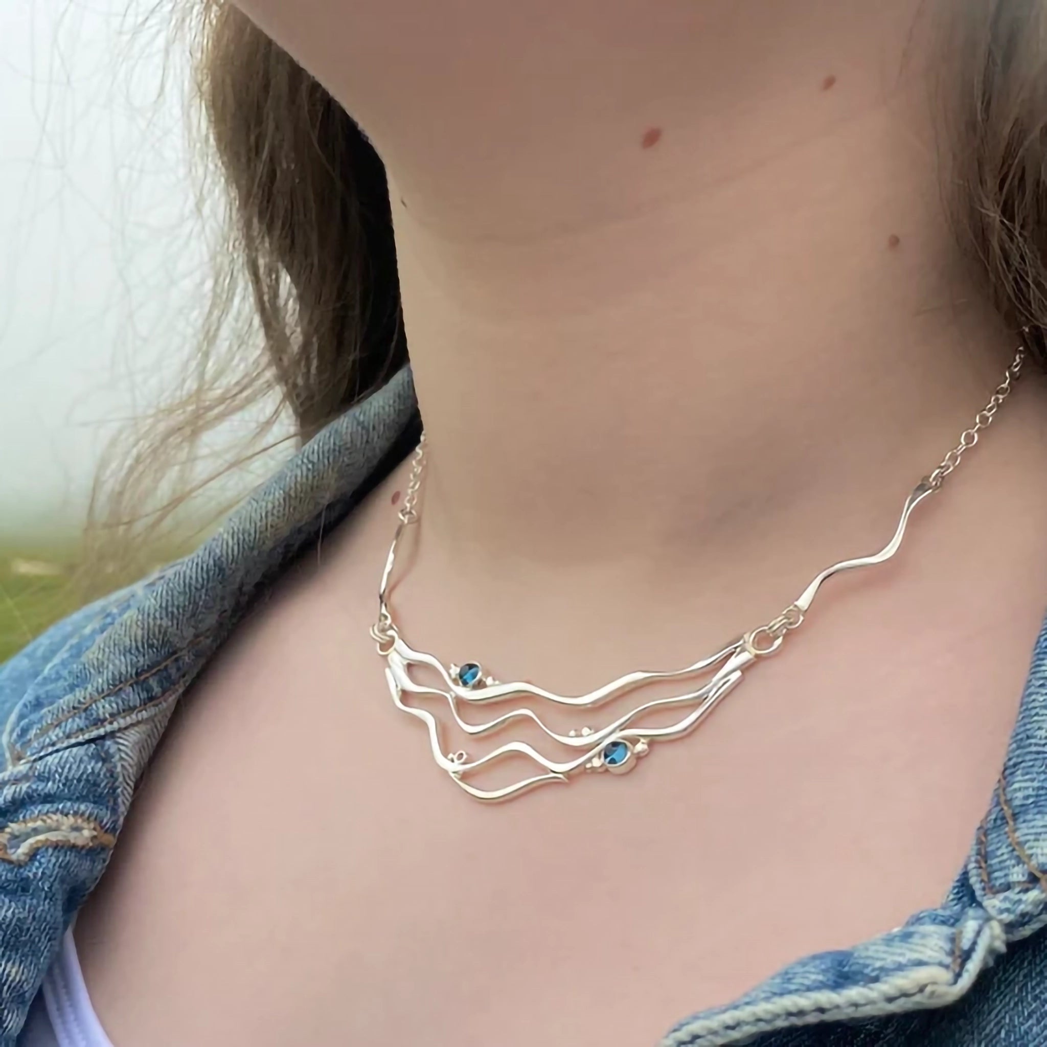 Model wearing a silver necklet featuring 4 ripple patterns and two blue topaz stones