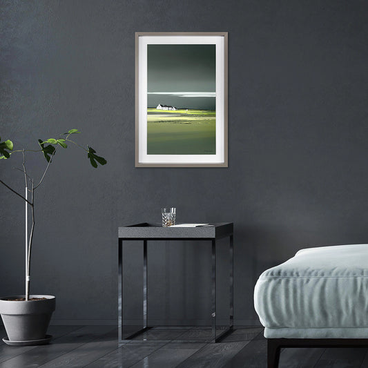 Framed print of a coastal scene in green and blue with a cluster of white cottages on the horizon hanging in dark room.