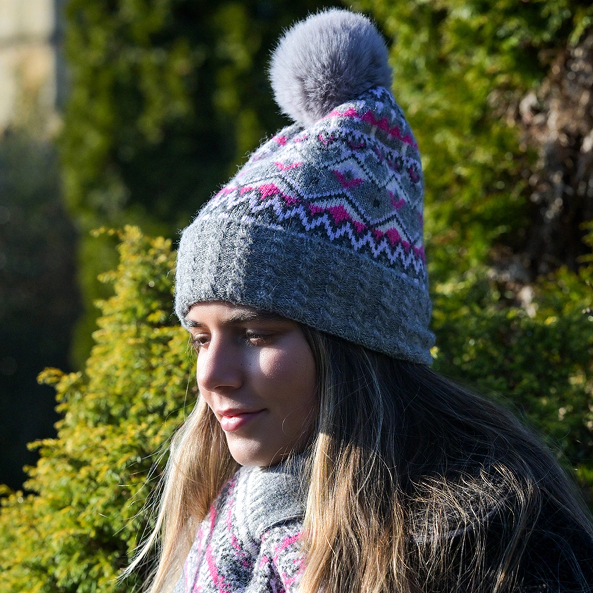 Model wearing knitted fair isle patterned hat in grey and pink with faux fur pompom and cable knit turn up
