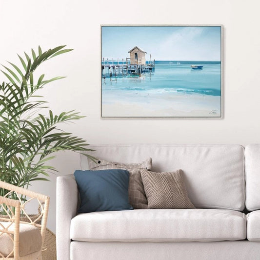 Framed canvas print with a wooden pier at the seaside and white boats in the distance hanging in a light room.