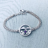 A silver bracelet with double chains and a round pendant with a Scottish thistle in front of hills and an amethyst stone