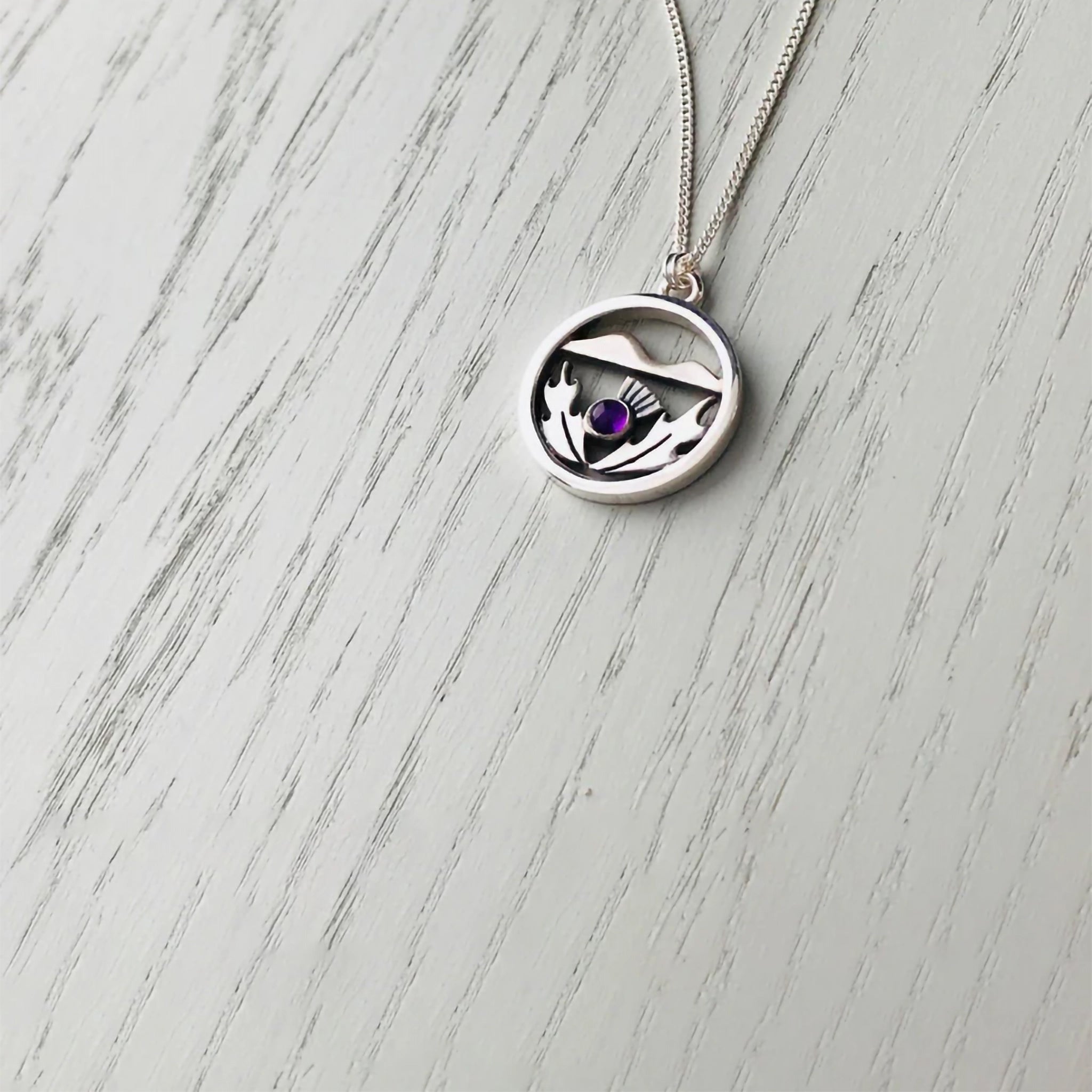 A round silver pendant with a Scottish thistle in front of hills and a purple amethyst stone