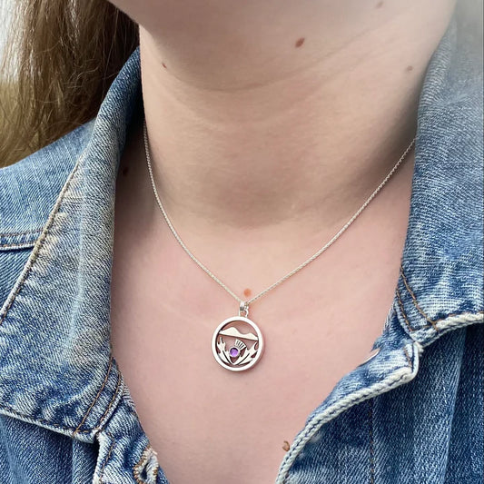 Model wearing a silver necklace with a pendant featuring a Scottish thistle in front of hill and a purple amethyst stone