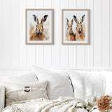 A wall art print of two hares in a neutral palette and wood effect frame hanging