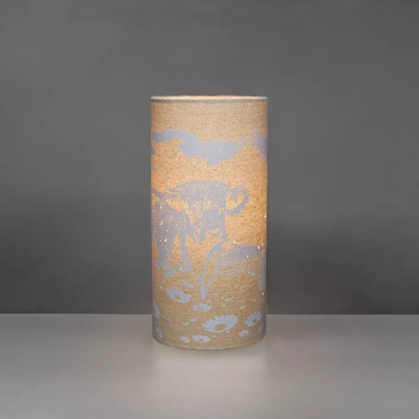 A linen look fabric table lamp with a Highland cow design and cut out details lit up