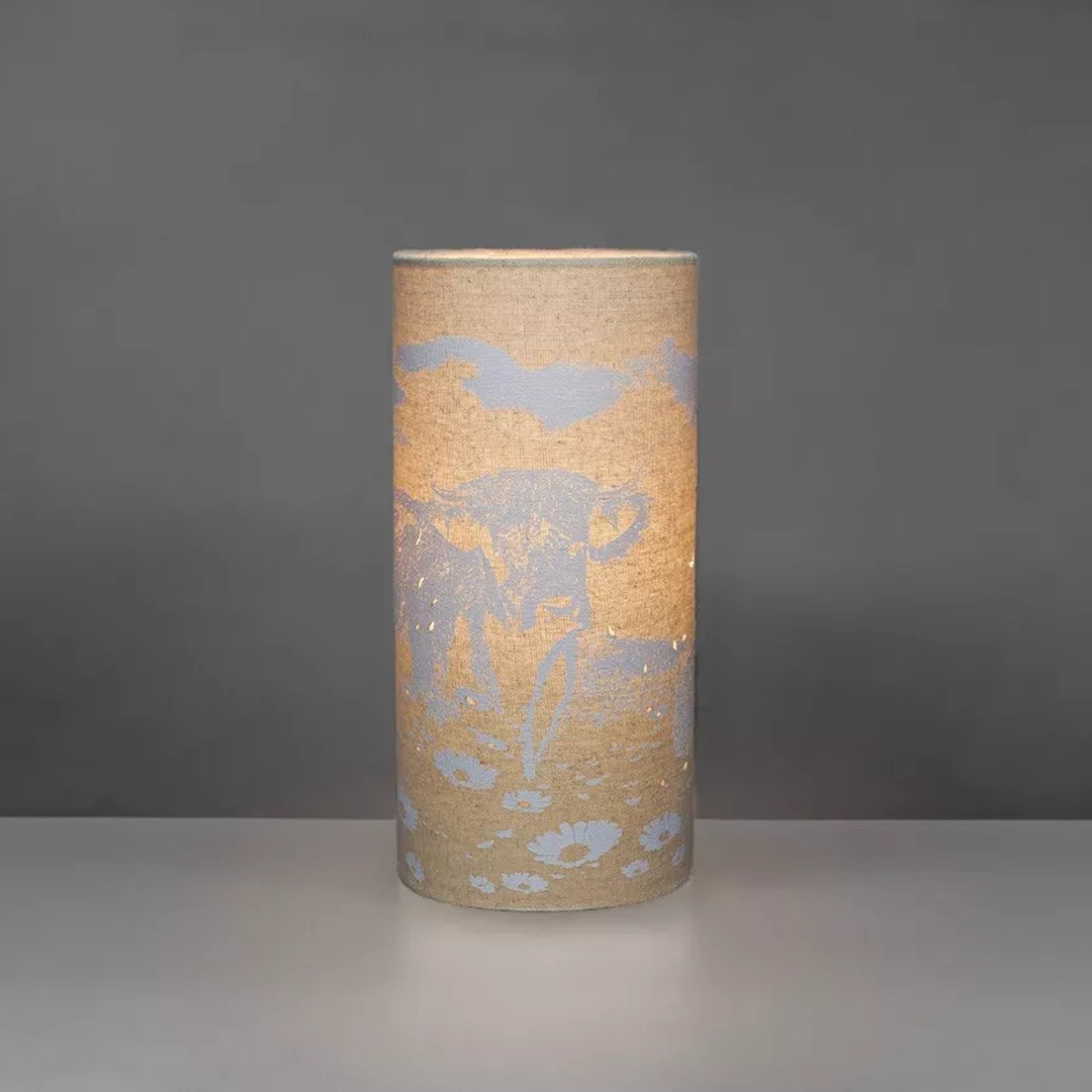 A linen look fabric table lamp with a Highland cow design and cut out details lit up