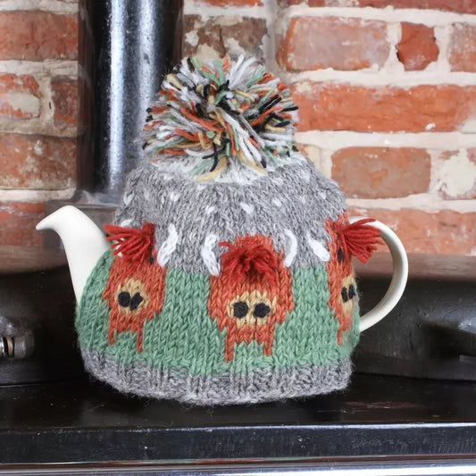 A knitted tea cosy featuring Highland cows and a pompom on a teapot